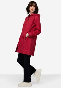 Veste imperméable rouge avec capuche, fermeture éclair sur le devant et poches latérales. Texture lisse et design mi-long, associée à un jean noir et des baskets.