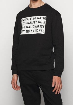 Person iført en sort sweatshirt med et hvidt rektangulært tryk, der gentager sætningen "NO NATIONALITY" med sorte store bogstaver.