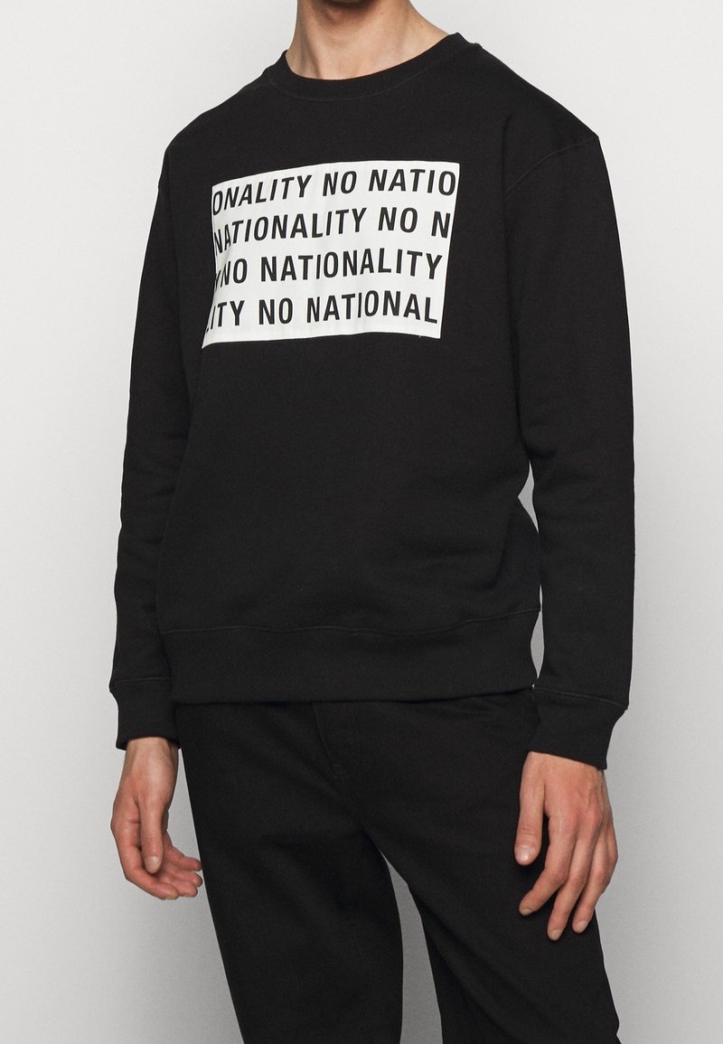Personne portant un sweat-shirt noir avec une impression rectangulaire blanche répétant la phrase « NO NATIONALITY » en lettres majuscules noires.