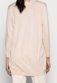 Kvinna som bär en ljusrosa långärmad tröja och krämfärgade byxor, sedd bakifrån med håret i en lös hästsvans.