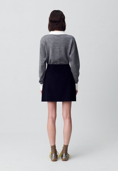 Suéter gris con cuello blanco, combinado con una falda negra. La modelo lleva calcetas marrones y zapatos plateados con cordones amarillos. Diseño minimalista.