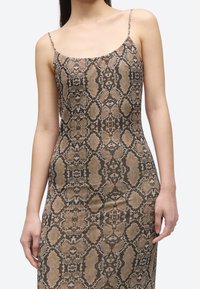 Naine, kes kannab varrukateta pruuni ja musta maduprintimisega kleiti, millel on õhukesed õlapaelad, ühtlase hele tausta ees.