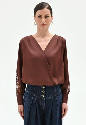 Donna che indossa una blusa marrone a maniche lunghe con dettagli in pizzo sulle maniche e jeans blu scuro a vita alta con pieghe e tre bottoni.