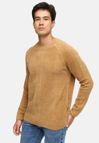 Mand iført en tekstureret kamelfarvet striktrøje og blå jeans, som står med den ene hånd bag ryggen mod en ensfarvet lys baggrund.