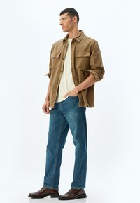 Camisa de pana marrón con dos bolsillos en el pecho, camiseta beige clara, jeans de mezclilla azules y botas de cuero marrón oscuro.