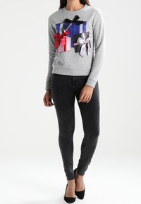 Felpa grigia con tre applicazioni a forma di scatola regalo strutturate in blu, rosso e rosa, ciascuna con fiocchi a contrasto. Abbinata a jeans skinny neri.