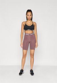 Must värvi spordibrassi, millel on valge logo, koos tumepunaste rattapükste ja valge vööga. Tüüpilised mustad spordijalatsid täiendavad riietust.