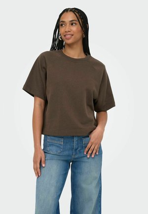 Femme aux cheveux tressés portant un haut marron à manches courtes et un jean bleu, souriante et regardant sur le côté avec une main dans la poche.