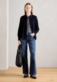 Chemise noire à boutons, haut texturé gris, jeans évasés bleus et escarpins noirs. Tient un sac noir à côtes. Sol en bois clair, fond neutre.