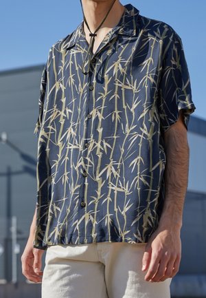 Chemise à manches courtes en tissu avec un fond bleu présentant un motif de bambou en vert clair. Dotée d'un boutonnage sur le devant et d'une coupe ample.