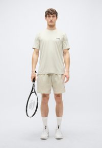 Bež športna majica s kratkimi rokavi in logotipom, kombinirana z ujemajočimi se kratkimi hlačami. Drži črno teniško lopar. Bele superge in nogavice.