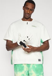 Λευκό τοπ με κοντά μανίκια και πράσινες πινελιές tie-dye, σε συνδυασμό με λευκά γάντια που φέρουν μαύρα μπαλώματα, τονίζοντας ένα casual αθλητικό στυλ.