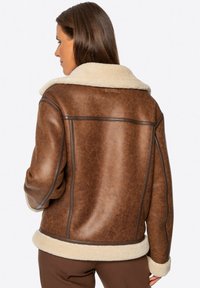 Braune Kunstlederjacke mit cremefarbigem Lammfell-Ausschnitt und -Manschetten. Sie hat eine lockere Passform und sichtbare Nähte.