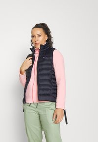 Marinblå pufferväst med hög krage och dragkedjefickor, lager på lager över en rosa zip-up tröja, matchat med ljusgröna byxor.