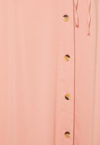 Vaaleanpunainen mekko, jossa on sileä pinta, nappiyksityiskohtia yhdellä sivulla ja solmittava vyötärö.