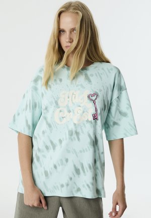 T-shirt surdimensionné vert menthe avec un motif tie-dye, présentant un grand graphique avec du texte et un détail de clé en forme de cœur, ainsi que des accents en strass.