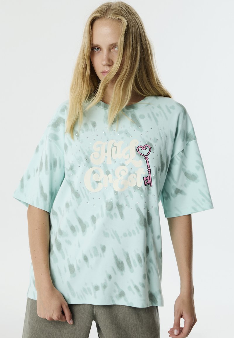 T-shirt de gran tamaño en verde menta con un patrón de tie-dye, que presenta un gráfico grande con texto y un detalle de llave en forma de corazón, además de acentos de strass.