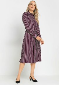 Abito lungo bordeaux con strisce bianche, dotato di colletto, maniche lunghe, vita con cintura e gonna svasata. Il materiale sembra liscio.