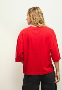 Červené bavlněné tričko s volným střihem, kulatým výstřihem a spadlými rameny. Obsahuje boční rozparky a rovný lem. Uvedený zadní pohled.