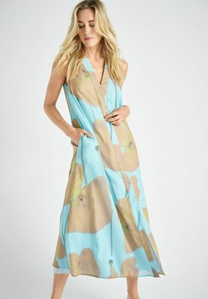 Blonde vrouw in een mouwloze blauwe jurk met grote beige bloemmotieven, handen in de zakken, staand tegen een effen lichte achtergrond.