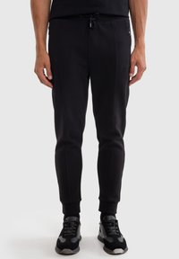 Pantalones de chándal negros con cintura ajustable, corte ajustado, bolsillos laterales con cremallera y puños acanalados, fabricados en una tela suave y texturizada.
