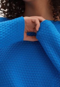 Niebieski sweter o fakturze z wzorem plastra miodu. Zbliżenie na tkany materiał i detal rękawa, pokazujące intensywny kolor i fakturę.