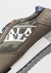 Gros plan d'une basket en daim marron, tissu camouflage vert, détail bleu marine, semelle intermédiaire blanche et semelle extérieure grise texturée.