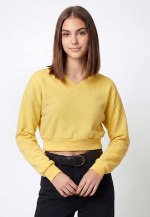Ung kvinde med glat brunt hår iført en gul cropped sweater og sorte bukser, står med hænderne løst foldet.