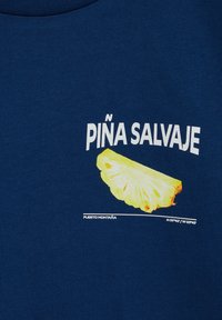 Žuta grafika kriške ananasa s tekstom "PIÑA SALVAJE" bijele boje na tamnoplavoj tkanini. Ispod se nalaze koordinate i "Puerto Montana".