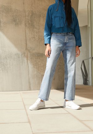 Person standing indoors wearing a blue cropped jacket, light blue high-waisted jeans, and white sneakers on tiled floor.