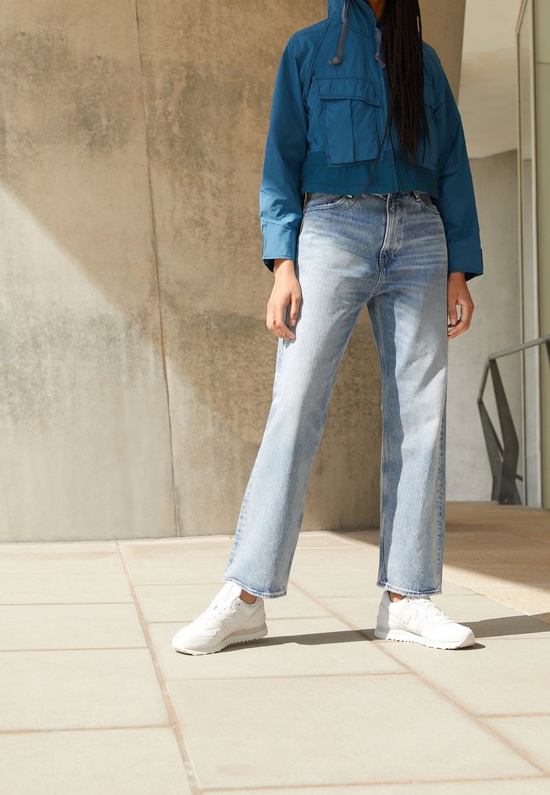 Personne debout à l'intérieur portant une veste bleue courte, un jean taille haute bleu clair et des baskets blanches sur un sol carrelé.