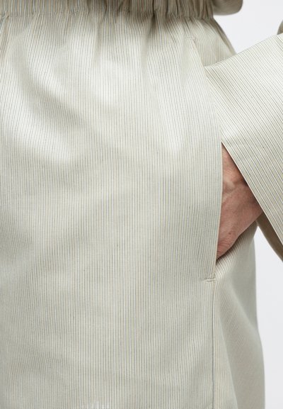 Tissu beige rayé avec une texture douce, présentant un design superposé, une poche visible et des accents bleus subtils dans les rayures.