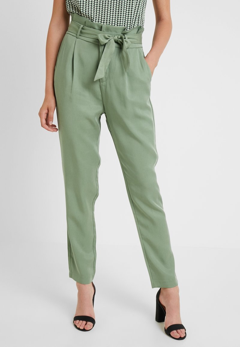 Un pantalon vert clair à taille haute, doté de plis et d'une ceinture à nouer. Texture lisse, jambes fuselées et poches latérales. Porté avec des talons noirs.