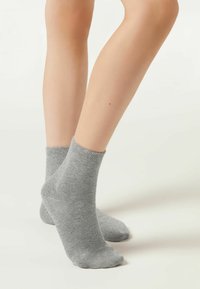 Calzini alla caviglia grigi in cotone con una texture liscia, bordo a costine e tallone rinforzato, mostrati su piedi nudi su uno sfondo chiaro.