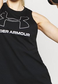 Person som lyfter en hantel, iklädd en svart ärmlös tröja från Under Armour, visar sin höjda muskulösa arm och axel.