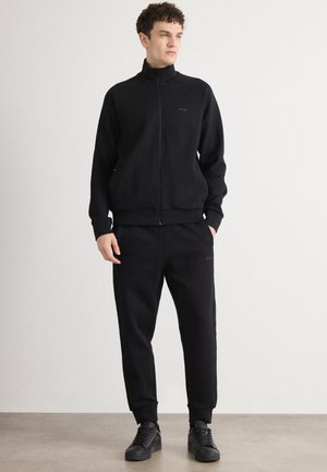 Chaqueta negra con cremallera y cuello alto, combinada con pantalones jogger negros. Ambos tienen una textura suave y un sutil detalle de logo. Usados con zapatos negros.