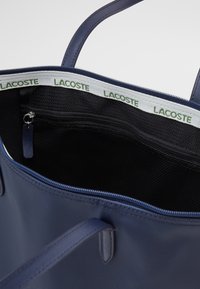Marinblå tote-väska med slitstarkt tyg, dragkedjestängning och svart insida. Har Lacoste-logotyp på en vit tygkant.