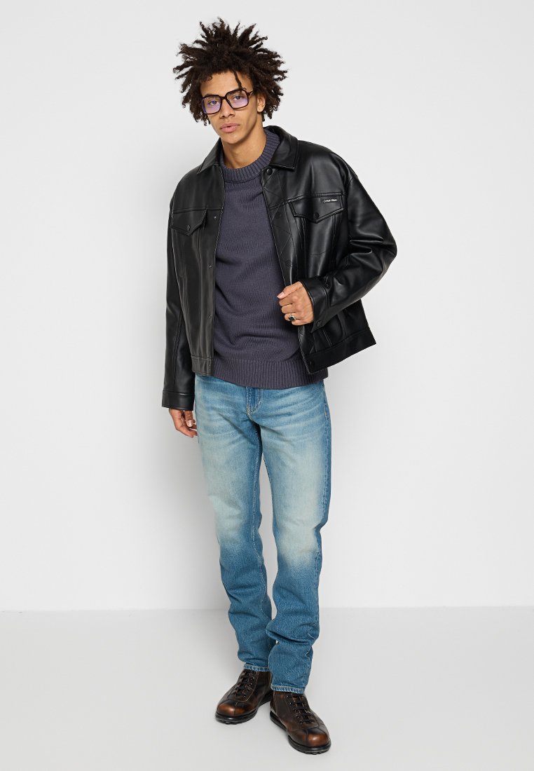 Giacca di pelle nera con due tasche frontali, indossata sopra un maglione a maglia grigio scuro. Jeans blu chiaro e stivali marroni stringati completano l'outfit.