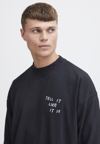 Black sweatshirt with a crew neckline, featuring white printed text "TELL IT LIKE IT IS" on the left chest area. Soft material, unstructured fit.