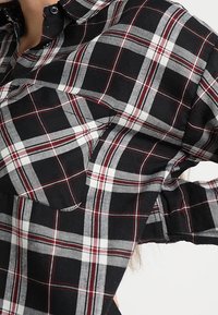 Black, white, and red plaid shirt with a front pocket and buttoned collar, worn by a person with long hair.