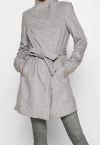 Persona che indossa un cappotto in lana grigia chiara con cintura e tasche con zip, abbinato a pantaloni grigi attillati su uno sfondo neutro.