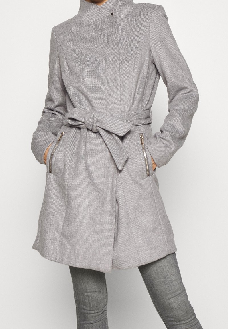 Persona che indossa un cappotto in lana grigia chiara con cintura e tasche con zip, abbinato a pantaloni grigi attillati su uno sfondo neutro.