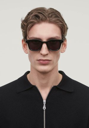 Black sunglasses with a rectangular frame, dark lenses, and subtle accents. Model wears a black zip-up sweater with a collared neck.