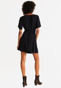 Vestido negro de manga corta con cintura ceñida y falda acampanada. Llevado con botas de tobillo. Tela suave con una textura blanda.