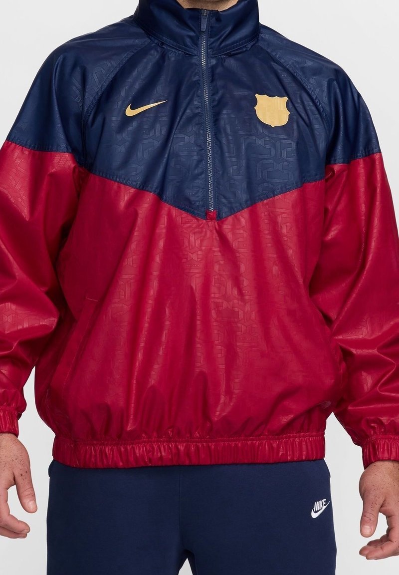 Cortavientos rojo y marino con media cremallera, puños elásticos, logo de Barcelona en dorado y patrón texturizado en la parte superior.