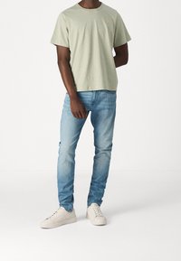 Ljusgrön bomullströja med korta ärmar och subtil logotyp. Är tillsammans med en tight passform av ljusblå jeans och beige sneakers. Enkel design.