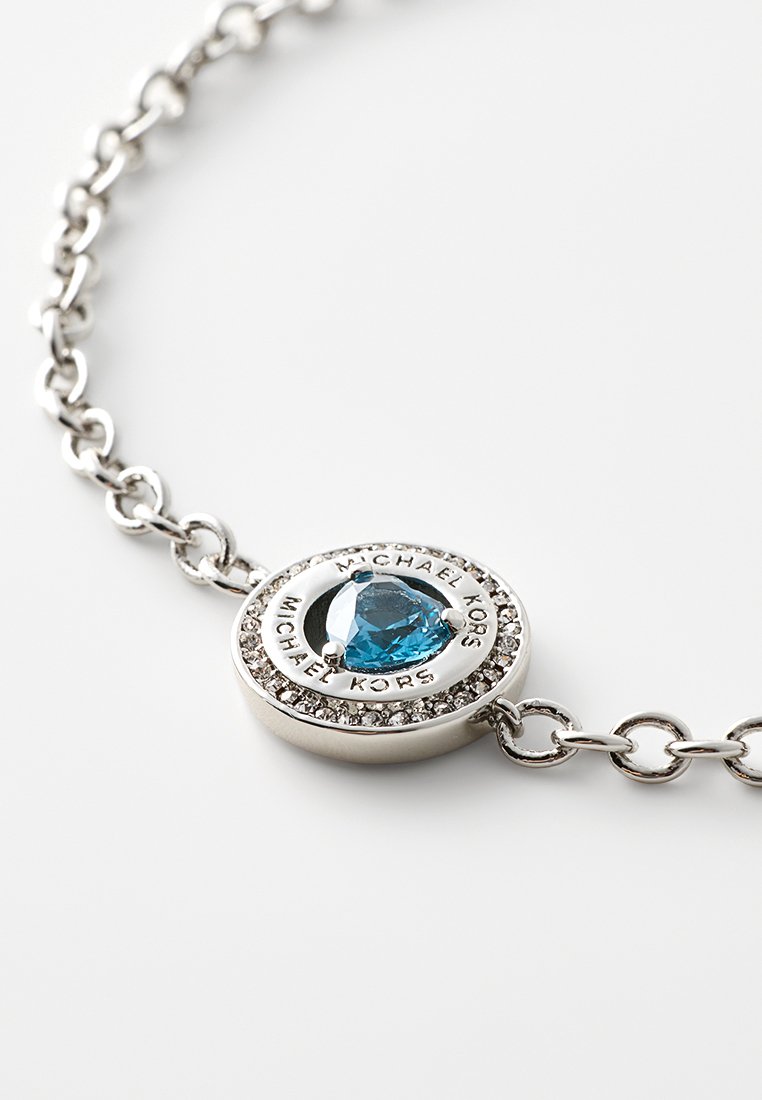 Srebrna bransoletka z łańcuchem i okrągłym wisiorkiem ozdobionym kryształami, który zawiera niebieski kamień w kształcie serca oraz wygrawerowany napis "MICHAEL KORS".