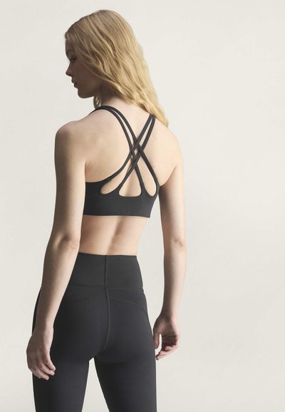 Donna con lunghi capelli biondi che indossa un reggiseno sportivo nero con spalline incrociate e leggings a vita alta, vista da dietro su sfondo semplice.