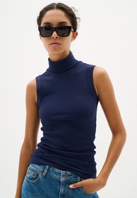 Top smanicato a collo alto a coste di colore blu navy con un design aderente, abbinato a jeans in denim. La modella indossa occhiali da sole rettangolari neri.