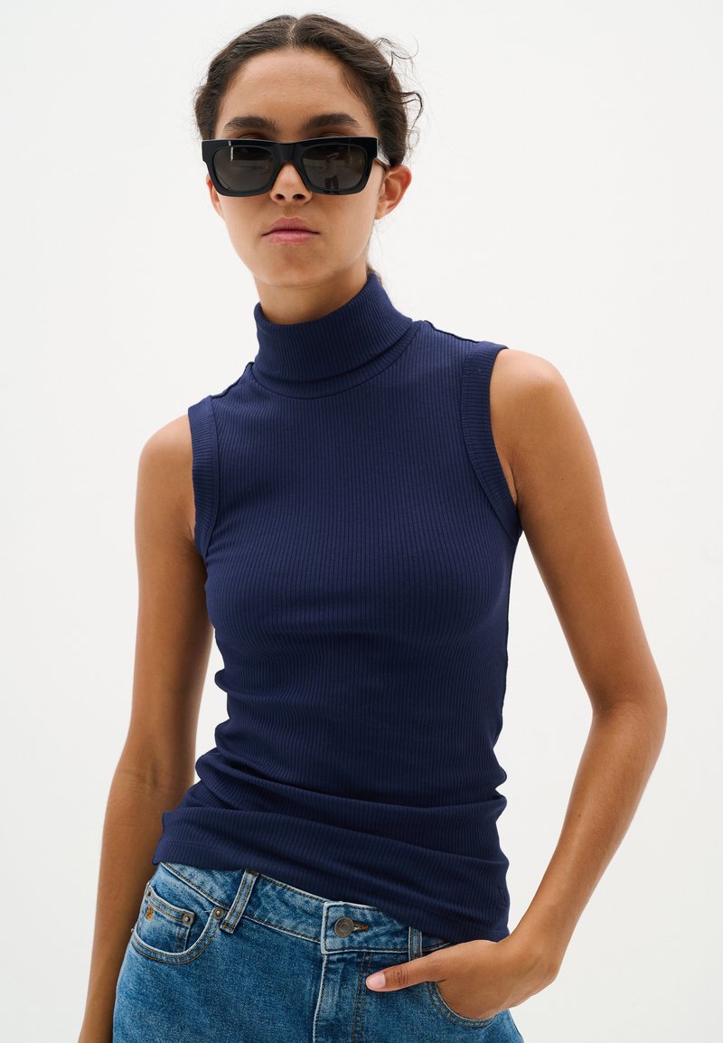 Top smanicato a collo alto a coste di colore blu navy con un design aderente, abbinato a jeans in denim. La modella indossa occhiali da sole rettangolari neri.